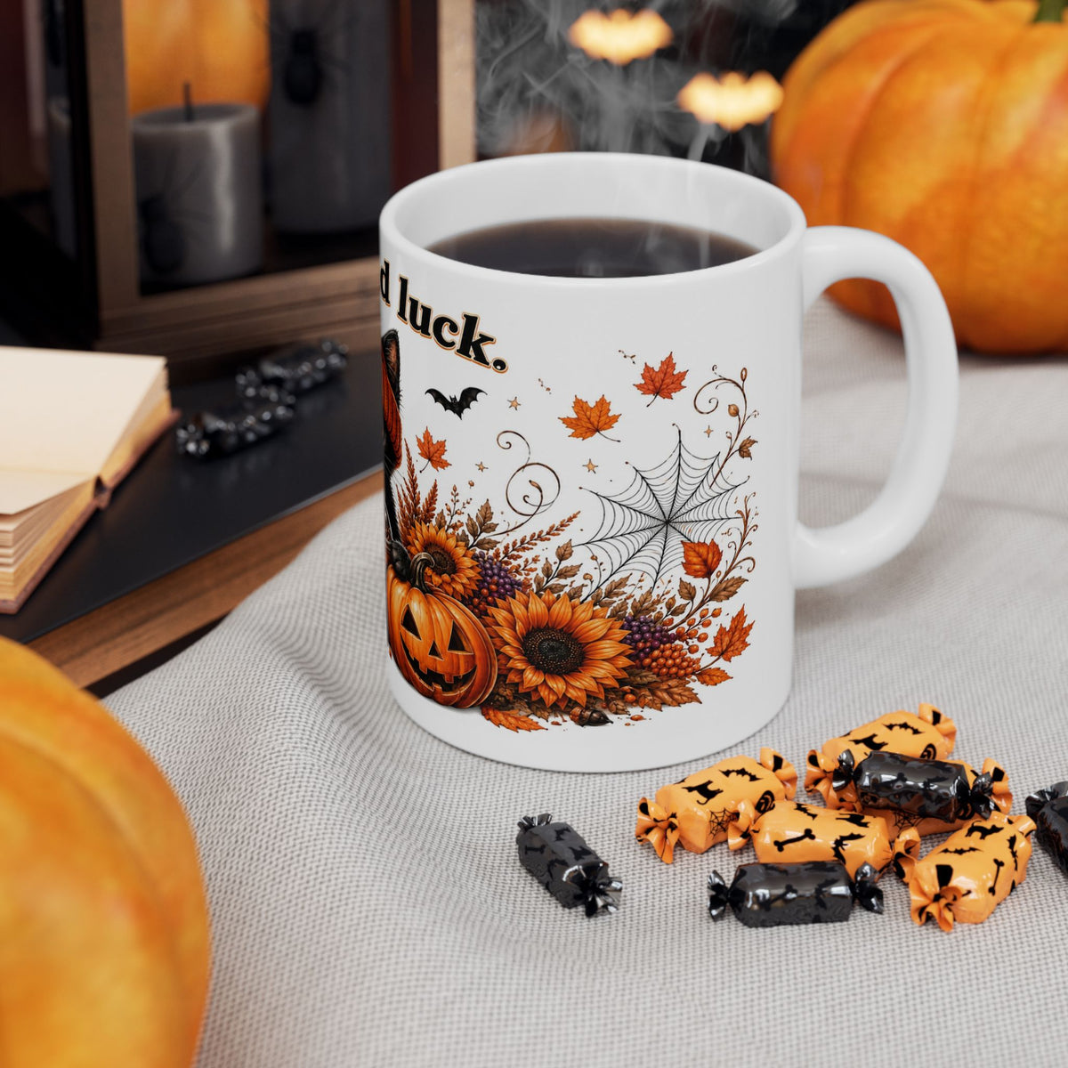 A white ceramic coffee mug with a Halloween-themed autumn print featuring jack-o'-lanterns, sunflowers, spiderwebs, and the text 'all luck.' It is filled with black coffee and styled on a light gray fabric tablecloth with pumpkins, candy, and candles in the background.