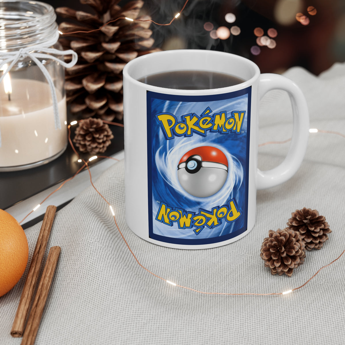 A white ceramic mug with a blue Pokémon graphic on the side, filled with a hot beverage and set on a light fabric surface with pinecones, a candle, cinnamon sticks, and fairy lights around it.